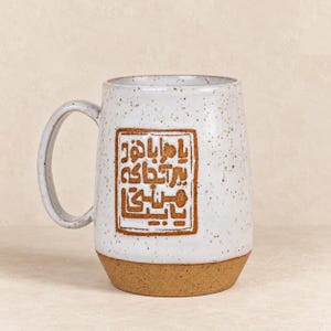May include: A speckled white ceramic mug with a large handle and a brown unglazed base. A rectangular brown stamp on the front features embossed Persian script text. The mug has a cylindrical shape with a smooth finish and visible dark brown flecks.