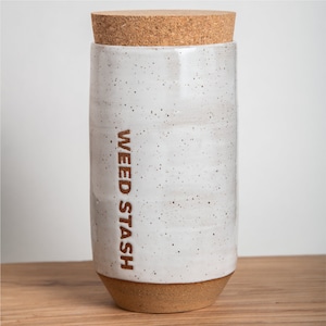May include: White ceramic jar with a cork lid and the words "WEED STASH" inscribed on the side. The jar has a speckled glaze and a brown base.