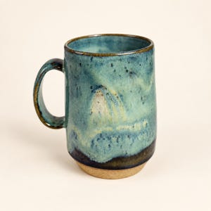 May include: A ceramic mug featuring a reactive glaze with shades of teal, blue, and green. The surface has a mottled, speckled texture with a darker blue base that transitions into a lighter, creamy tone. The bottom rim is unglazed, showing tan clay.