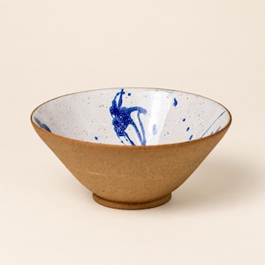 Op de afbeelding: Een keramische kom met een natuurlijke, gespikkelde beige buitenkant en een witte binnenkant met een levendig blauw verfspatpatroon. De kom heeft een brede, open rand en een kleine, ronde voet, wat een contrast vormt met de aardse textuur.