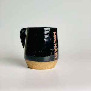 May include: Black ceramic mug with a tan rim and the name "MICHAEL" written in gold on the side.