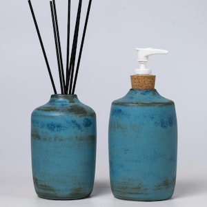 May include: A teal ceramic set comprising a bottle with a white pump and a vase with black reeds. The bottle has a cork stopper. The set has a rustic, handmade appearance. The set is suitable for the bathroom.