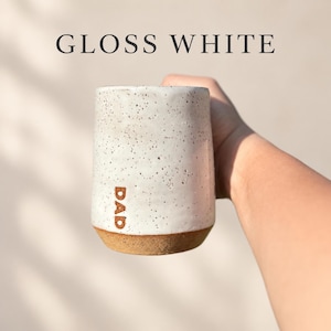 May include: A cylindrical ceramic mug with a glossy white speckled finish and a natural brown clay base. The word DAD is stamped vertically in brown letters on the side. The text GLOSS WHITE appears at the top of the image above the mug.
