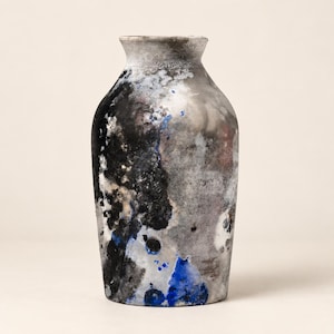 Puede incluir: Un jarrón de cerámica con cuerpo ancho y cónico y cuello estrecho. La superficie presenta un acabado abstracto y moteado en tonos negro, gris carbón, blanco y azul vibrante, creando una apariencia texturizada y desgastada en todo el exterior.