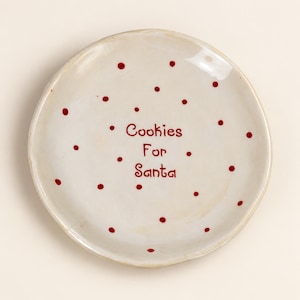 May include: A round, off-white ceramic plate with an uneven, hand-formed edge. The surface is decorated with small, scattered red polka dots and features the red text "Cookies For Santa" printed in the centre in a simple, rounded font style.