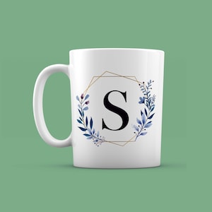 Peut inclure: Mug en céramique blanche avec un cadre géométrique doré et un motif floral bleu. La lettre "S" est au centre du cadre.