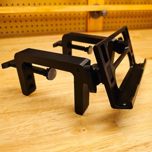 May include: Black 3D printed clamp-style phone holder with a built-in stand. The holder has a screw-type adjustment mechanism and a textured surface.