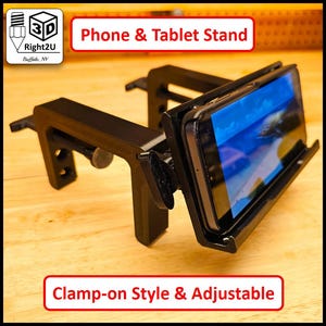 Puede incluir: Soporte negro para teléfono y tableta con estilo de abrazadera. El soporte es ajustable y sostiene una tableta. El soporte tiene el texto "Phone & Tablet Stand" y "Clamp-on Style & Adjustable". El soporte está sobre una superficie de madera.