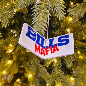 Op de afbeelding: Een witte kerstboomversiering met de woorden "BILLS MAFIA" in blauwe en rode letters. De versiering hangt aan een groene kerstboom met kleine, warmwitte lichtjes.