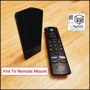 Op de afbeelding: Een zwarte 3D-geprinte houder voor een Fire TV-afstandsbediening. De houder is ontworpen om de afstandsbediening verticaal te houden. De afstandsbediening is zwart met een zilveren knop en een blauw "firetv"-logo.