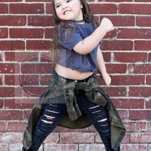 Pu&ograve; includere: Una bambina che indossa una maglietta grigia corta, jeans neri strappati e una giacca mimetica. Indossa stivali neri da combattimento e ha un fiocco verde nei capelli. &Egrave; in piedi davanti a un muro di mattoni.