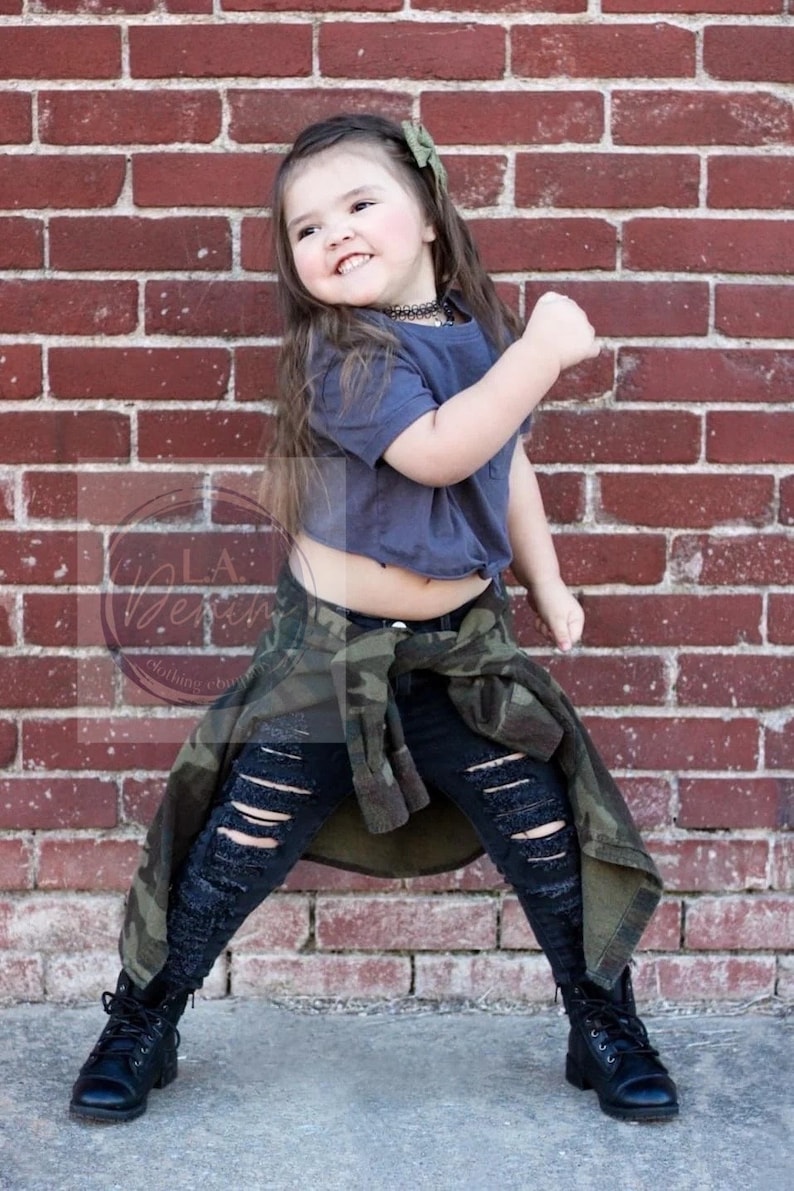 Pu&ograve; includere: Una bambina che indossa una maglietta grigia corta, jeans neri strappati e una giacca mimetica. Indossa stivali neri da combattimento e ha un fiocco verde nei capelli. &Egrave; in piedi davanti a un muro di mattoni.