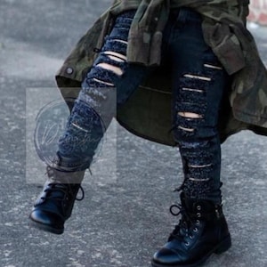 Pu&ograve; includere: Jeans neri strappati con bordi sfilacciati e stivali neri da combattimento. I jeans sono indossati da una persona che indossa una giacca mimetica.