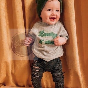 Pu&ograve; includere: Un bambino piccolo che indossa un berretto verde, una maglietta grigia con la scritta "Lucky & free" in verde, jeans neri strappati e scarpe da ginnastica nere e bianche.