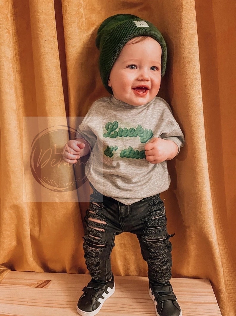 Pu&ograve; includere: Un bambino piccolo che indossa un berretto verde, una maglietta grigia con la scritta "Lucky & free" in verde, jeans neri strappati e scarpe da ginnastica nere e bianche.