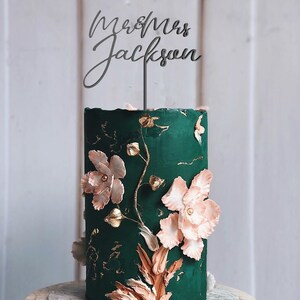 Op de afbeelding: Een donkergroene bruidstaart met bloemversieringen en gouden accenten. De taart is versierd met grote, lichtroze bloemen en een taarttopper met de tekst "Mr & Mrs Jackson". De taart staat op een marmeren taartstandaard.
