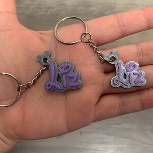 May include: Two silver keyrings with the name "Liz" written in purple on each charm. The keyrings are connected by a silver chain.