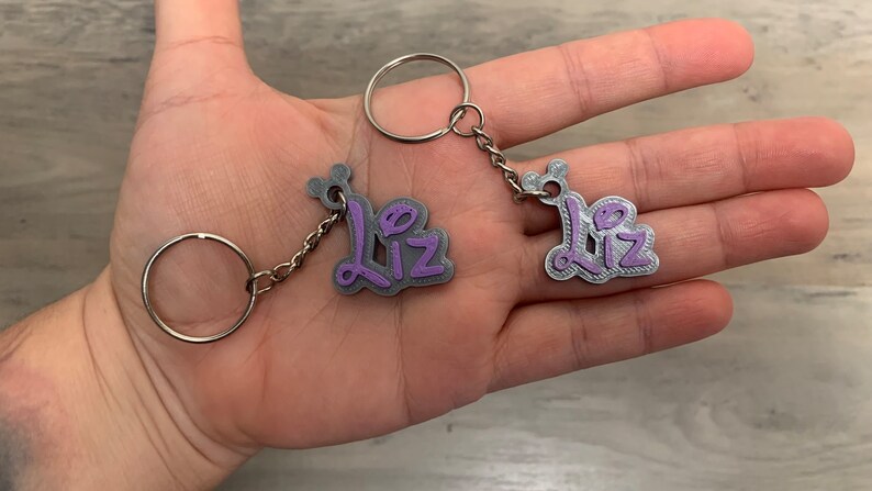 May include: Two silver keyrings with the name "Liz" written in purple on each charm. The keyrings are connected by a silver chain.
