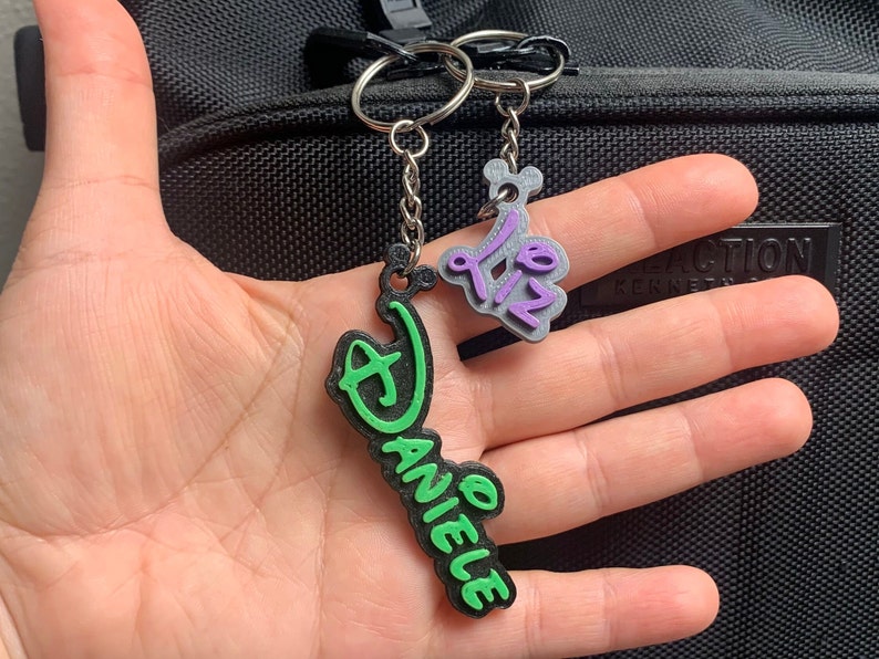 May include: Two keychains with personalised names. One keychain is green with the name "Danielle" in black lettering. The other keychain is purple with the name "Liz" in white lettering.