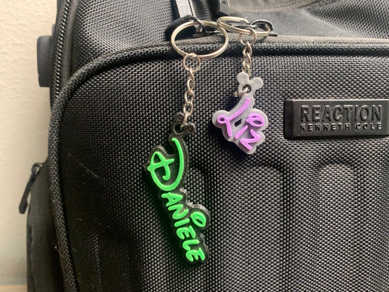 May include: Two keychains with personalised names. The keychain on the left is green and says "Danielle". The keychain on the right is purple and says "Liz".