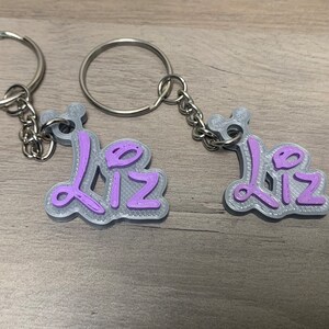 May include: Two silver keyrings with the name "Liz" written in purple on each. The keyrings are on a wooden surface.