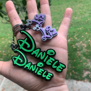 May include: A green and black keychain with the name "Daniele" written in a stylised font. There is a smaller purple keychain with the name "Liz" written in a stylised font.