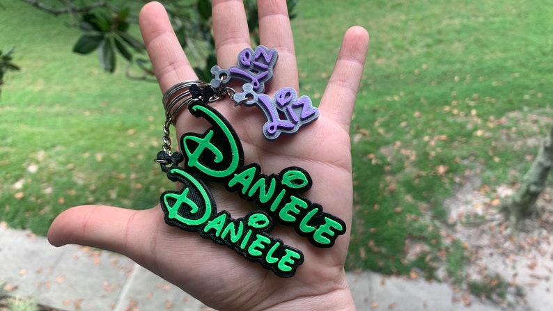 May include: A green and black keychain with the name "Daniele" written in a stylised font. There is a smaller purple keychain with the name "Liz" written in a stylised font.
