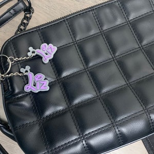May include: A black quilted crossbody bag with a silver chain strap and two purple keychains with the name "Liz" on them.
