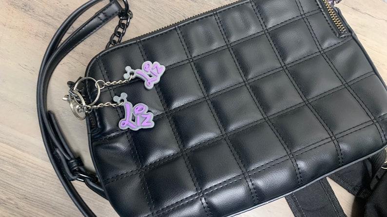 May include: A black quilted crossbody bag with a silver chain strap and two purple keychains with the name "Liz" on them.