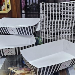 Könnte beinhalten: Rechteckige weiße Papier-Lebensmittelbehälter mit schwarzen vertikalen Streifen. Die Behälter haben eine weiße Innenseite und kleine Griffaussparungen. Die Behälter tragen die Buchstaben "PR" auf der Vorderseite. Ein Stapel ähnlicher Behälter ist ebenfalls sichtbar.