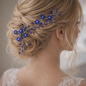 Pode incluir: Um acessório de cabelo composto por um ramo de arame prateado adornado com pequenas contas azuis em forma de flor e minúsculas pérolas brancas. Os ramos flexíveis estendem-se, criando um arranjo floral delicado preso no cabelo loiro.