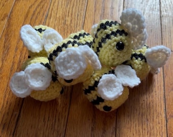 May include: A cluster of handmade crochet bees. Each bee features a yellow body with black stripes, white wings, and black eyes. The bees are arranged on a wooden surface, showcasing their handcrafted details.