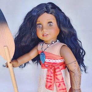 Puede incluir: Una mu&ntilde;eca de piel morena con el pelo largo y negro rizado que lleva un vestido marr&oacute;n y naranja con una faja roja. La mu&ntilde;eca sostiene una pala de madera y lleva un collar con una joya azul. La mu&ntilde;eca tiene un tatuaje en el brazo.