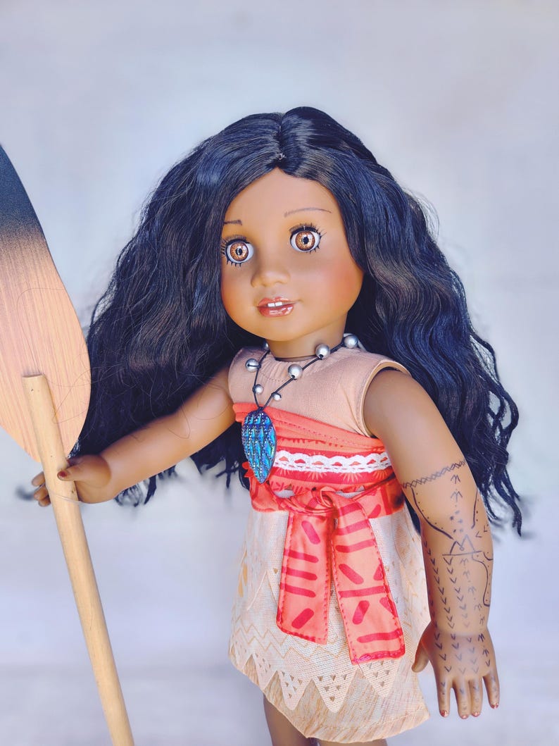 Puede incluir: Una mu&ntilde;eca de piel morena con el pelo largo y negro rizado que lleva un vestido marr&oacute;n y naranja con una faja roja. La mu&ntilde;eca sostiene una pala de madera y lleva un collar con una joya azul. La mu&ntilde;eca tiene un tatuaje en el brazo.