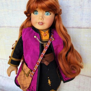 May include: A red-haired doll wearing a black dress with a purple vest, a brown belt, and a brown leather bag. The doll has blue eyes and is wearing a gold necklace.