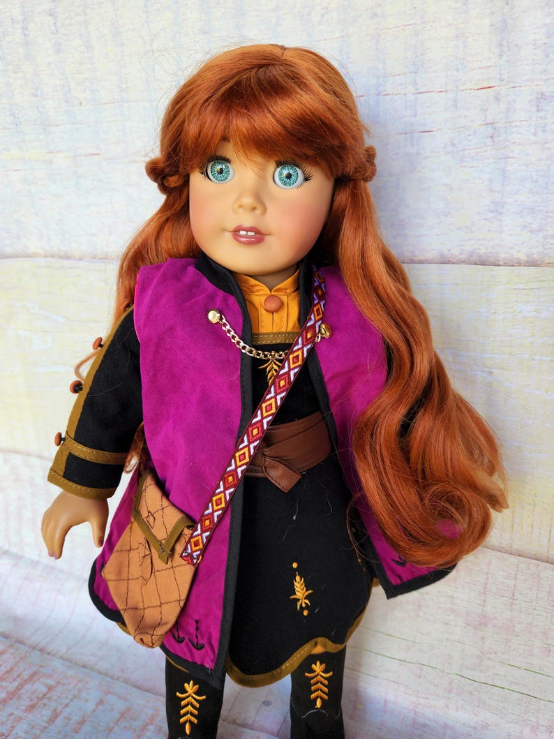 May include: A red-haired doll wearing a black dress with a purple vest, a brown belt, and a brown leather bag. The doll has blue eyes and is wearing a gold necklace.