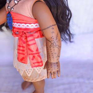 Puede incluir: Una mu&ntilde;eca de piel morena que lleva un vestido rosa y naranja con ribete blanco y un collar azul. La mu&ntilde;eca tiene el pelo largo y negro y un tatuaje negro en el brazo.
