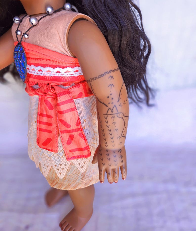 Puede incluir: Una mu&ntilde;eca de piel morena que lleva un vestido rosa y naranja con ribete blanco y un collar azul. La mu&ntilde;eca tiene el pelo largo y negro y un tatuaje negro en el brazo.