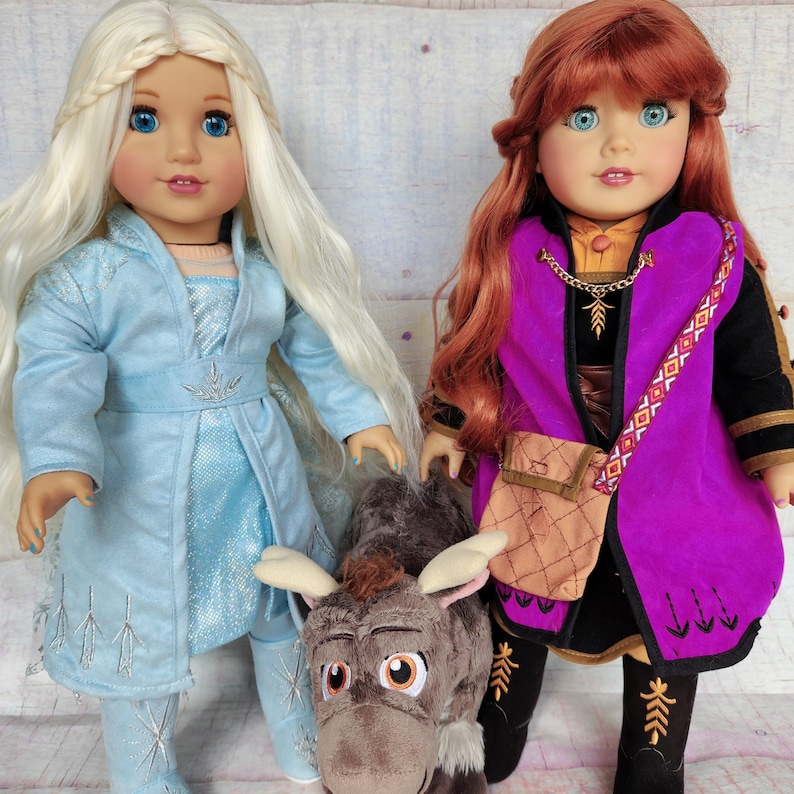 May include: Two dolls dressed as Elsa and Anna from the movie Frozen. The doll dressed as Elsa has long blonde hair and is wearing a light blue dress with a snowflake design. The doll dressed as Anna has long red hair and is wearing a purple dress with a brown belt and a snowflake design. The dolls are standing next to a brown plush toy that looks like Sven the reindeer.