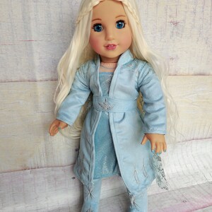 May include: A doll wearing a light blue dress and matching coat with silver snowflake designs. The doll has long blonde hair and blue eyes.