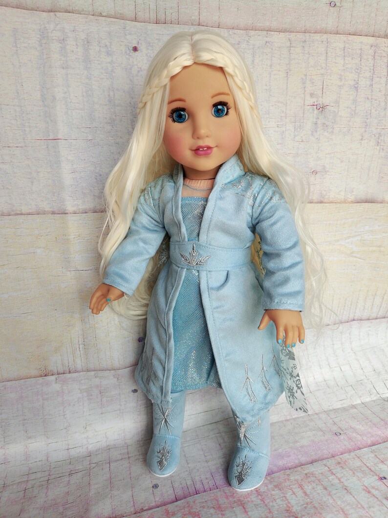 May include: A doll wearing a light blue dress and matching coat with silver snowflake designs. The doll has long blonde hair and blue eyes.