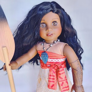 Puede incluir: Una mu&ntilde;eca de piel morena con el pelo largo y negro rizado que lleva un vestido rojo y tostado con un collar azul y turquesa. La mu&ntilde;eca sostiene una pala de madera.