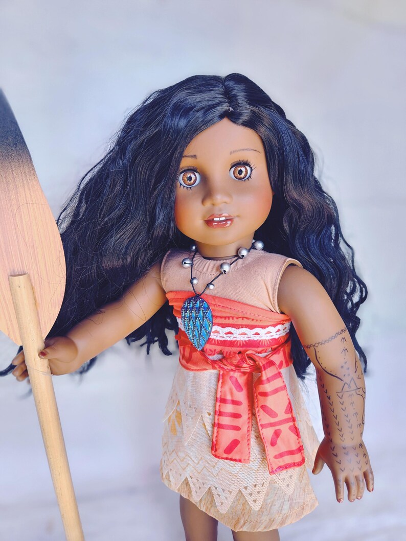 Puede incluir: Una mu&ntilde;eca de piel morena con el pelo largo y negro rizado que lleva un vestido rojo y tostado con un collar azul y turquesa. La mu&ntilde;eca sostiene una pala de madera.