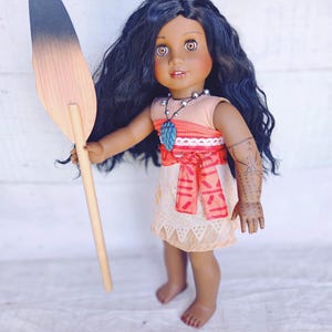 Puede incluir: Una mu&ntilde;eca de piel morena con el pelo largo y rizado oscuro lleva un vestido de color melocot&oacute;n con una faja de estampado rojo y blanco. Est&aacute; sosteniendo una pala de madera con un degradado de negro a marr&oacute;n. La mu&ntilde;eca lleva un collar con un colgante azul y verde.