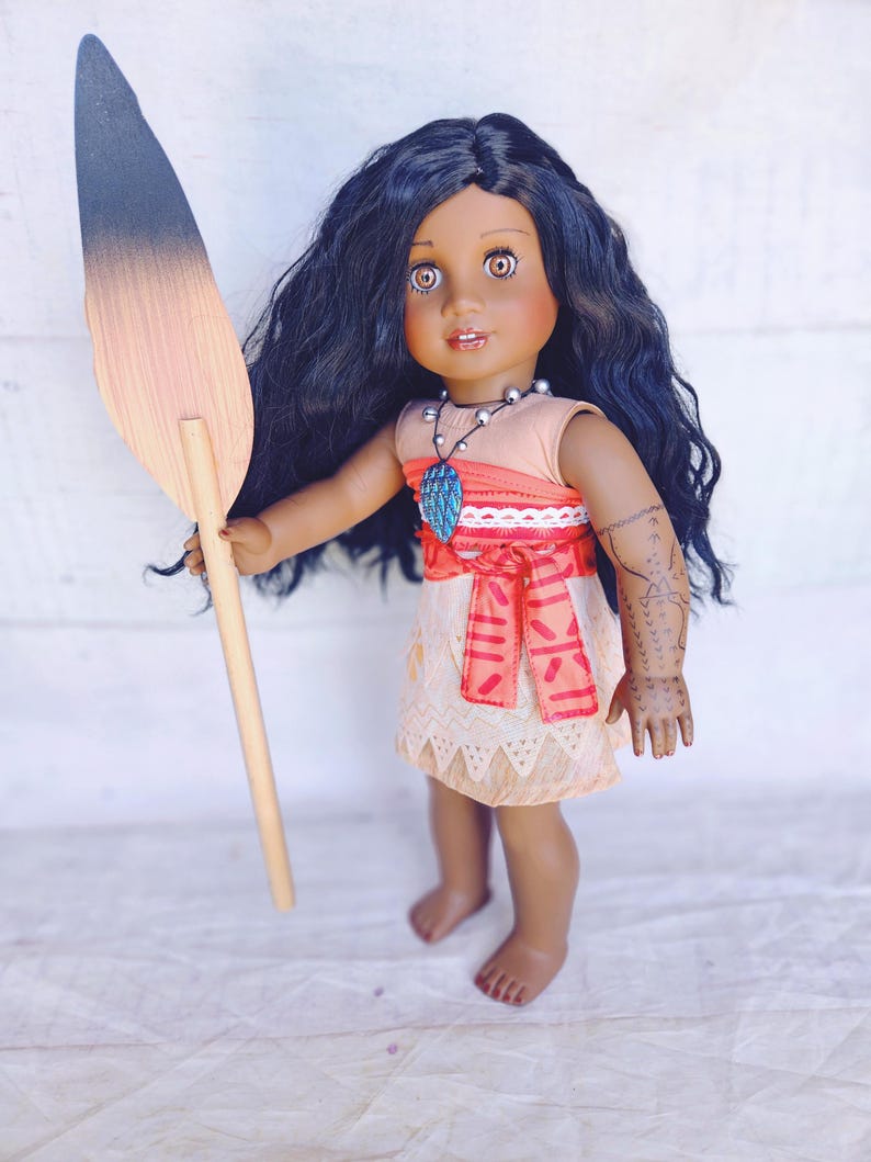 Puede incluir: Una mu&ntilde;eca de piel morena con el pelo largo y rizado oscuro lleva un vestido de color melocot&oacute;n con una faja de estampado rojo y blanco. Est&aacute; sosteniendo una pala de madera con un degradado de negro a marr&oacute;n. La mu&ntilde;eca lleva un collar con un colgante azul y verde.