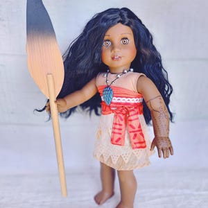 Puede incluir: Una mu&ntilde;eca de piel morena con el pelo largo y negro rizado lleva un vestido rojo y naranja con ribete de encaje blanco. Est&aacute; sosteniendo una lanza de madera con una punta negra y marr&oacute;n. La mu&ntilde;eca lleva un collar con un colgante azul y tiene un tatuaje negro en el brazo.