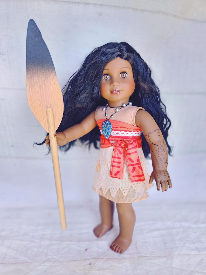 Puede incluir: Una mu&ntilde;eca de piel morena con el pelo largo y negro rizado lleva un vestido rojo y naranja con ribete de encaje blanco. Est&aacute; sosteniendo una lanza de madera con una punta negra y marr&oacute;n. La mu&ntilde;eca lleva un collar con un colgante azul y tiene un tatuaje negro en el brazo.