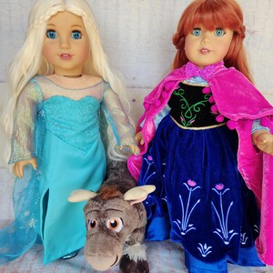 May include: Two dolls dressed as Elsa and Anna from the movie Frozen. Elsa is wearing a blue and white dress with a long blonde braid. Anna is wearing a blue and pink dress with a red cape and a long red braid. A brown plush reindeer toy is in front of the dolls.
