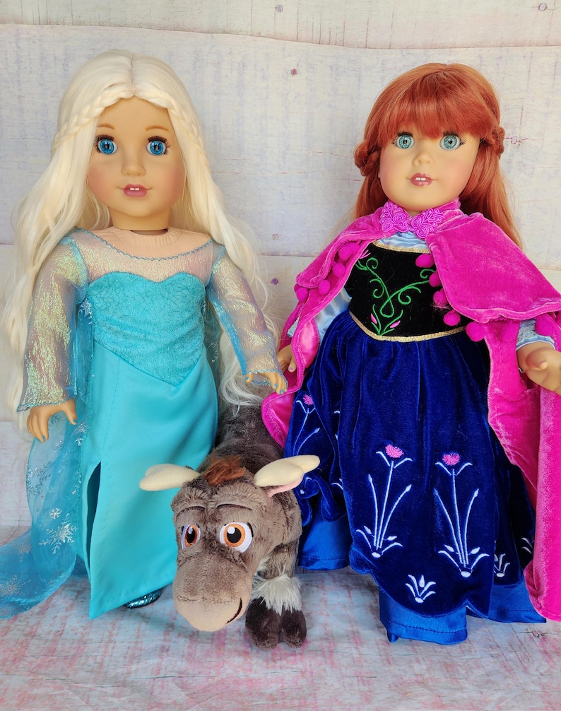May include: Two dolls dressed as Elsa and Anna from the movie Frozen. Elsa is wearing a blue and white dress with a long blonde braid. Anna is wearing a blue and pink dress with a red cape and a long red braid. A brown plush reindeer toy is in front of the dolls.