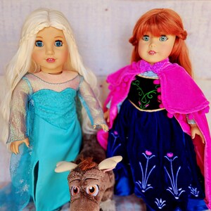 May include: Two dolls dressed as Elsa and Anna from the movie Frozen. The doll on the left is wearing a light blue dress with a sparkly bodice and a long blonde braid. The doll on the right is wearing a dark blue dress with a pink cape and a red wig. The dolls are standing next to a brown plush toy.